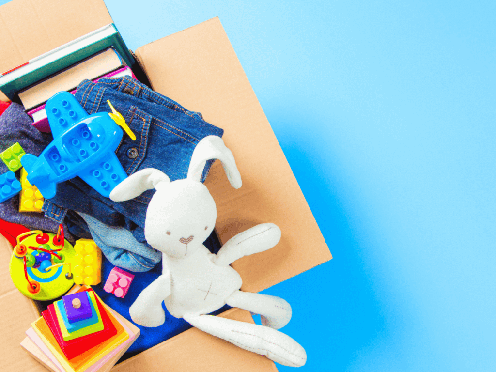a cardboard box of various children's toys and clothes including a toy plane and a white bunny. Background is sky blue.