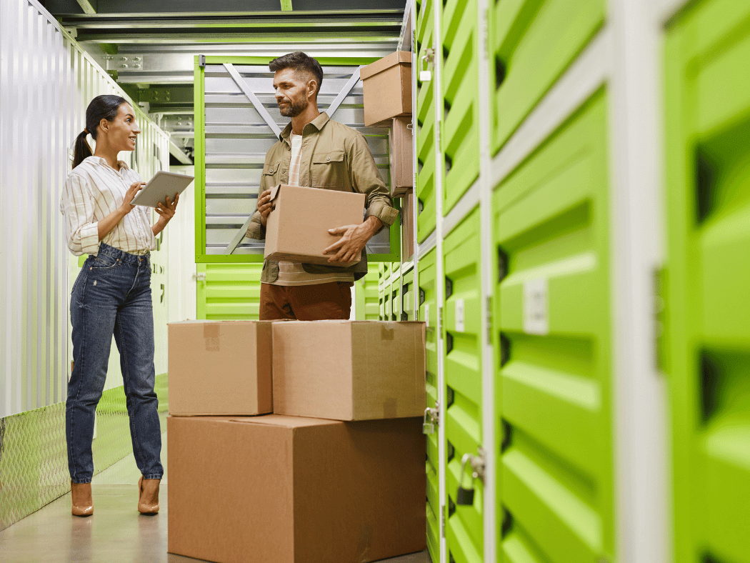 A man and a woman stand in front of a small storage unit with a checklist and several boxes.