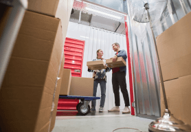 a couple holding boxes are standing outside their well organised long term storage unit