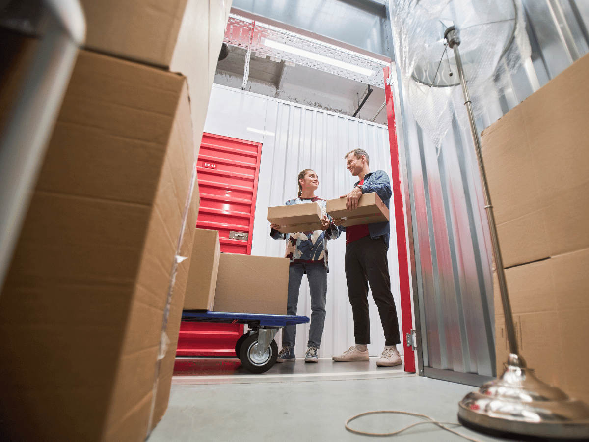 a couple holding boxes are standing outside their well organised long term storage unit