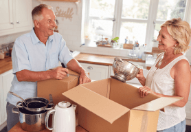 An older couple are in the process of downsizing. They are in their kitchen, smiling as they pack kitchen items into boxes.