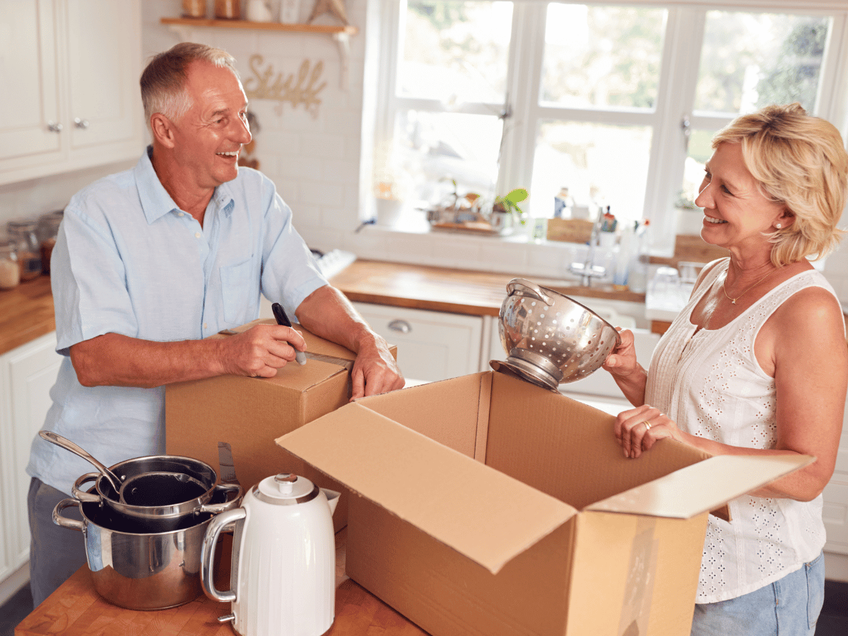 An older couple are in the process of downsizing. They are in their kitchen, smiling as they pack kitchen items into boxes.