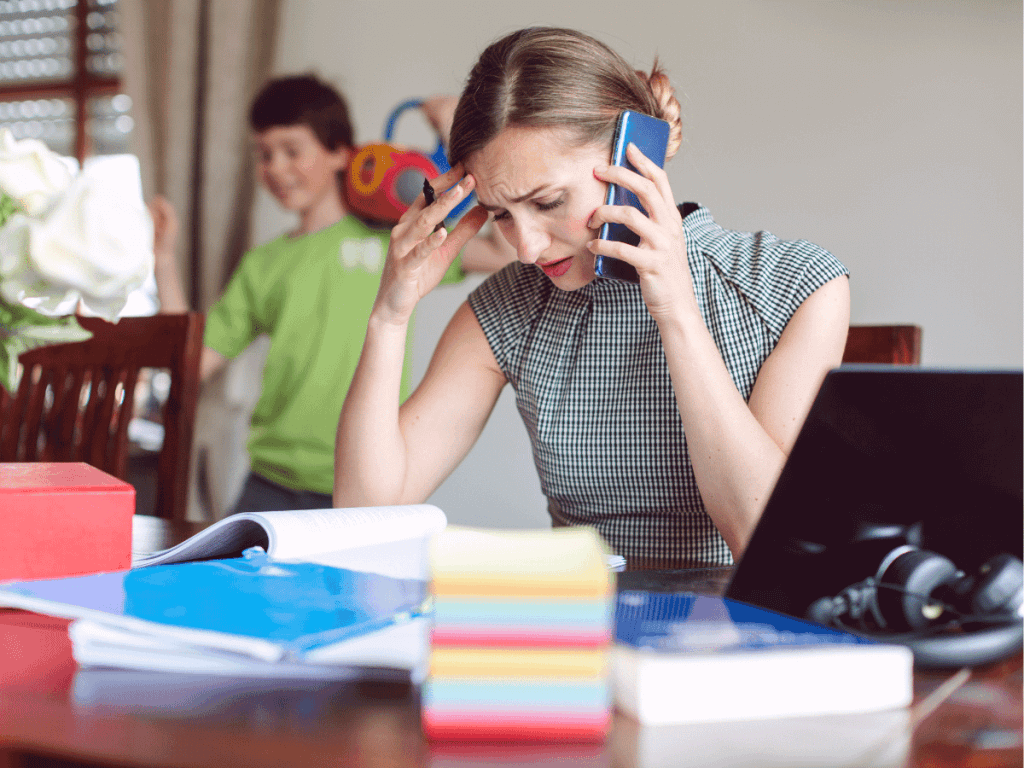 A woman is trying to make a telephone call in her new business at home. The desk is cluttered with notebooks and a child is playing in the background.