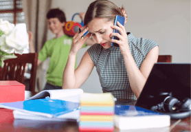 A woman is trying to make a telephone call in her new business at home. The desk is cluttered with notebooks and a child is playing in the background.